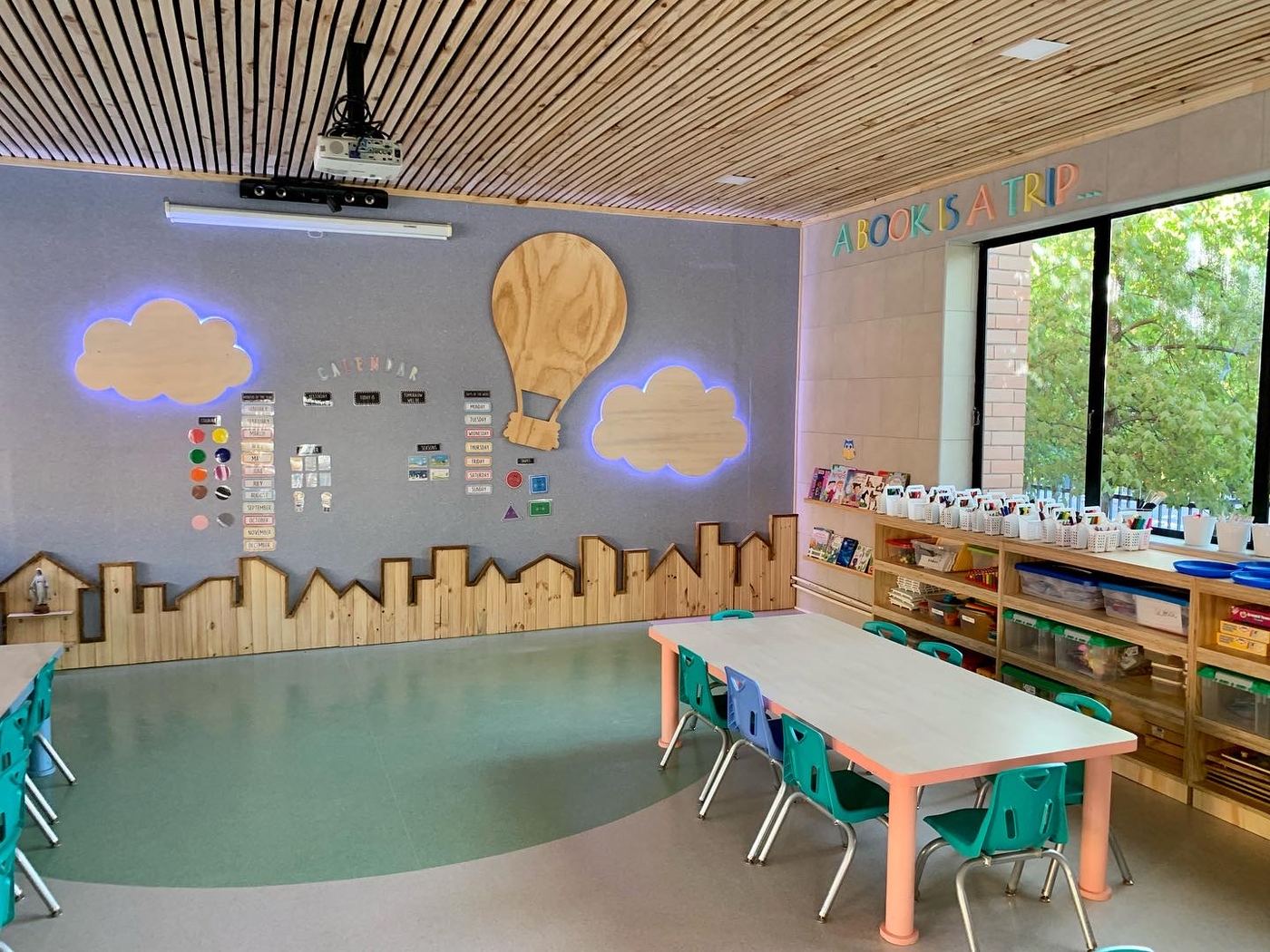 Sala con cielo de listones de madera, ventanal hacia los árboles y muebles de terciado